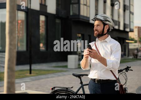 Beau jeune homme avec vélo et téléphone mobile debout dans la rue après la journée de travail. Portrait d'un homme d'affaires à barbe confiant dans un casque à l'aide d'un téléphone cellulaire, Banque D'Images