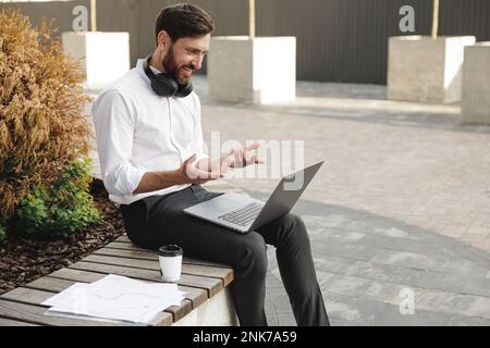Sympathique indépendant homme souriant et gestant pendant la conversation vidéo sur ordinateur portable. Homme plein de joie avec un casque sur le cou assis à l'extérieur sur un banc avec Banque D'Images
