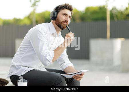 Un employé de bureau sérieux regarde avec soin tout en tenant le stylet et l'ordinateur portable entre les mains. Homme aux cheveux foncés en chemise blanche, assis dans un quartier urbain pendant que pla Banque D'Images