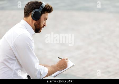 Homme barbu en chemise blanche générant des idées créatives et écrivant dans un carnet. Homme caucasien indépendant écoutant de la musique dans des écouteurs sans fil pendant que W Banque D'Images