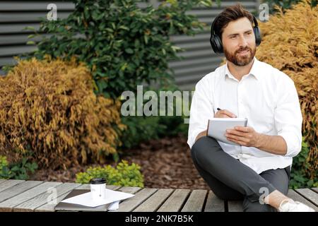 Homme d'affaires plein de joie dans les écouteurs pensant à un nouveau projet et écrivant des idées dans un ordinateur portable. Homme souriant assis sur le banc dans la zone urbaine avec ordinateur portable, faites Banque D'Images