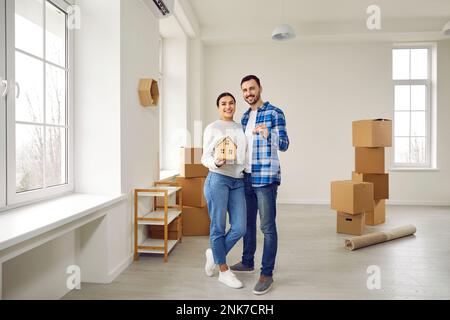 Photo pleine grandeur d'un couple heureux marié tenant les clés de leur nouvel appartement spacieux et moderne et maquette de maison de jouet. Un couple familial regarde dans l'appareil photo Banque D'Images