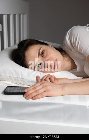 Une femme de 30 ans se trouve dans un lit avec un smartphone près d'un oreiller, addiction téléphonique Banque D'Images