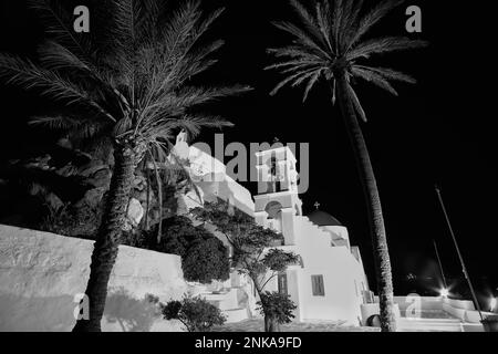 L'église orthodoxe blanchie à la chaux illuminée de Panagia Gremniotissa à iOS Grèce, entourée de palmiers la nuit en noir et blanc Banque D'Images