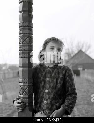 Comté de Suceava, République socialiste de Roumanie, env. 1980. Fille assise sur le porche de sa maison par un pilier en bois magnifiquement sculpté. Banque D'Images