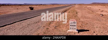 Route vers Agadir. Jalon indiquant Agadir dans une zone désertique du Maroc. en arrière-plan, une petite silhouette d'un homme sur une moto. Banque D'Images