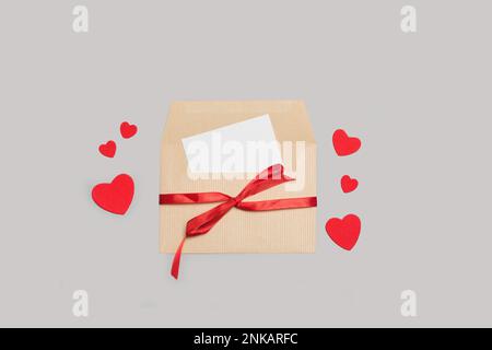 Un papier blanc vierge à l'intérieur d'une enveloppe marron attaché avec un ruban rouge sur un fond gris avec des coeurs en papier rouge Banque D'Images