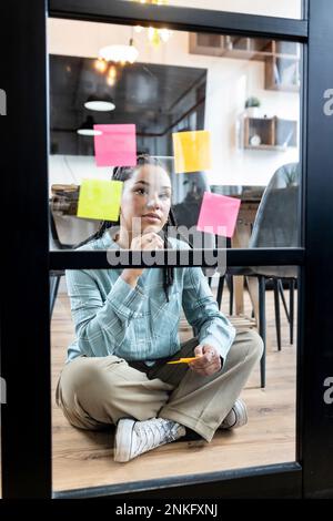 Femme d'affaires contemplative lisant des notes d'adhésifs sur la porte en verre dans le bureau Banque D'Images