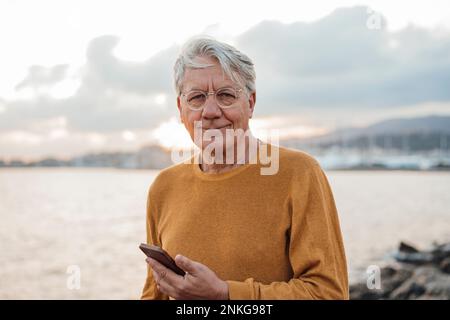 Homme senior souriant debout avec un téléphone portable en face de la mer Banque D'Images