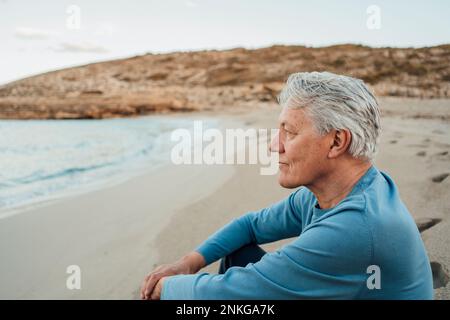 Homme senior attentionné, assis sur la plage au coucher du soleil Banque D'Images