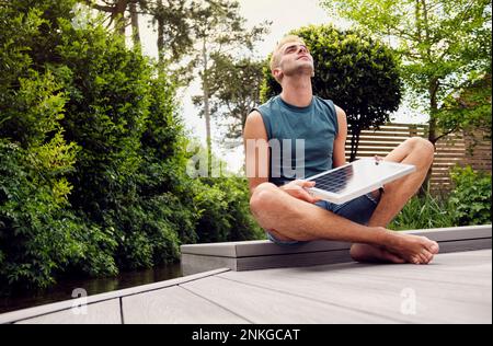 Jeune homme tenant un panneau solaire regardant vers le haut assis à pieds croisés sur le patio Banque D'Images