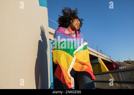 Jeune femme joyeuse enveloppée de drapeau arc-en-ciel en appréciant le soleil Banque D'Images