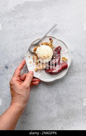 Main de femme tenant une assiette de poires au vin rouge avec de la crème glacée et du granola Banque D'Images