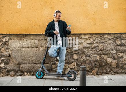 Homme heureux utilisant un smartphone debout avec un scooter électrique en face du mur Banque D'Images