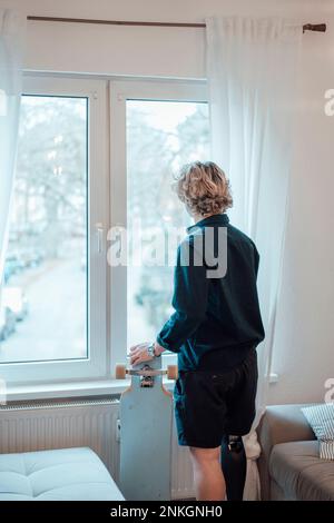 Jeune homme debout avec un skateboard devant la fenêtre à la maison Banque D'Images