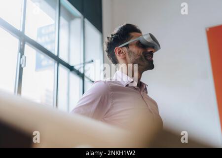 Homme d'affaires mûr souriant portant des lunettes VR au bureau Banque D'Images