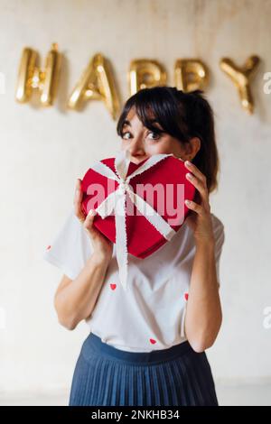 Femme avec boîte cadeau en forme de coeur debout devant le mur Banque D'Images