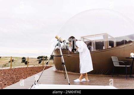 Jeune femme portant un peignoir regardant à travers le télescope par tente dôme Banque D'Images