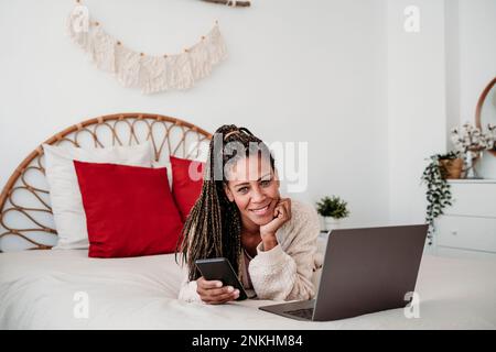 Un pigiste souriant allongé sur le lit avec un ordinateur portable et un smartphone à la maison Banque D'Images