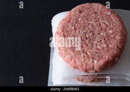 Hamburgers de bœuf crus empilés sur une planche à découper en ardoise, séparés par du papier sulfurisé Banque D'Images