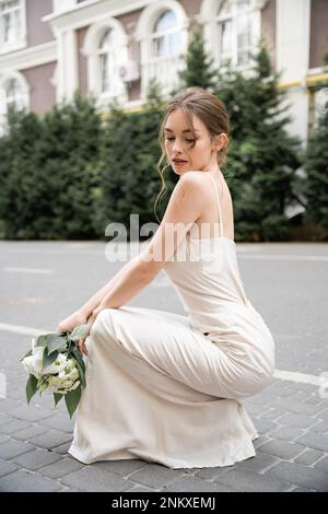 jeune mariée en robe de mariage tenant bouquet de fleurs tout en étant assis à l'extérieur, image de stock Banque D'Images