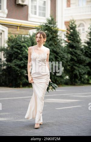 pleine longueur de jolie mariée en robe blanche tenant le bouquet de mariage et la marche sur la rue, image de stock Banque D'Images