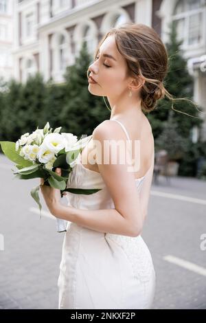 jeune mariée en robe blanche debout avec les yeux fermés et tenant le bouquet de mariage, image de stock Banque D'Images