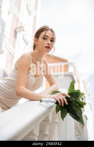 vue à faible angle de la superbe mariée dans la robe de mariage tenant le bouquet et regardant l'appareil photo près de la maison, image de stock Banque D'Images