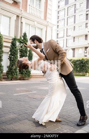 pleine longueur de mariée joyeuse dans la robe de mariage tenant le bouquet et la flexion près de marié, image de stock Banque D'Images