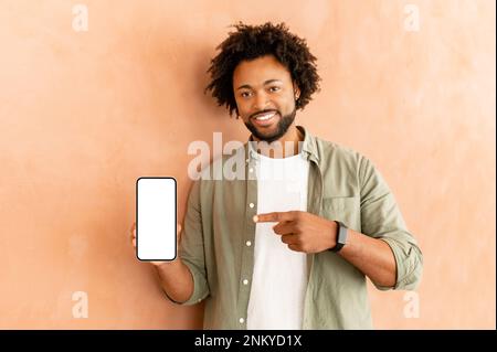 Homme afro-américain debout isolé sur le fond du mur, présentant une nouvelle application, pointant avec les mains sur le téléphone mobile avec espace blanc pour la publicité, image maquette Banque D'Images