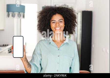 Bonne jeune femme avec une coiffure afro montrant un smartphone avec un écran vide avec l'intérieur de l'appartement sur fond, femme de maison gaie recommandant l'application mobile pour le shopping ou quik livraison à la porte Banque D'Images