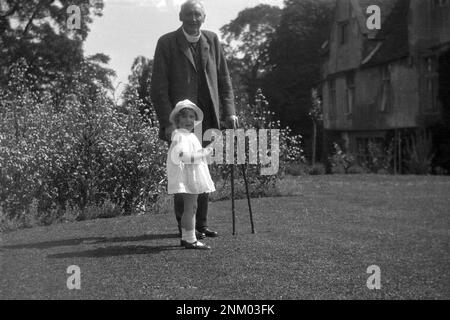 1931, historique, l'été et dehors sur une pelouse, un vicaire âgé avec bâtons de marche avec petite fille, sa petite-fille, Angleterre, Royaume-Uni. Le vicaire porte un costume en laine de trois pièces. Banque D'Images