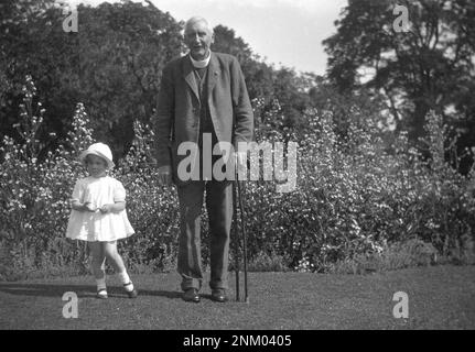 1931, historique, l'été et dehors sur une pelouse, un vicaire âgé avec bâtons de marche avec petite fille, sa petite-fille, Angleterre, Royaume-Uni. Le vicaire porte un costume en laine de trois pièces avec chaîne de montre. Banque D'Images