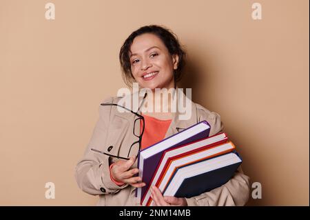 Femme d'âge moyen enseignante dans un établissement d'enseignement secondaire, souriant à la caméra, portant une pile de livres sur fond beige Banque D'Images