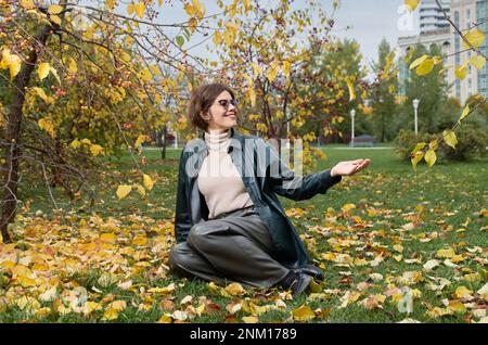Une femme vêtue de cuir est assise sur le sol au milieu de feuilles jaunes dans un parc d'automne Banque D'Images