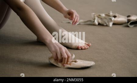 Une belle danseuse de ballet se trouve sur le sol près de pointe au studio de danse et met des chaussures de ballet avant la leçon de chorégraphie. La ballerine se prépare à la performance. École de danse classique. Pieds rapprochés. Banque D'Images