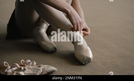 La ballerine adulte est assise sur le sol au studio de danse et met des chaussures pointe avant la leçon de chorégraphie. Un danseur magnifique se prépare à la performance ou à l'entraînement. École de danse classique. Pieds rapprochés. Banque D'Images