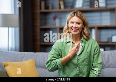 Souriante senior belle jeune femme assise sur un canapé à la maison devant l'appareil photo et parlant en langue des signes. Banque D'Images