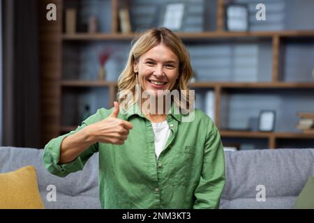 Souriant senior belle jeune femme assise à la maison sur le canapé devant l'appareil photo, montrant le doigt super, tout est ok. Banque D'Images
