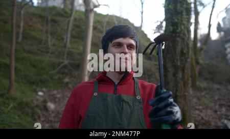 Fermier ou jardinier jeune homme en robe rouge avec tablier vert, paire regardant la caméra et souriant, montrant son concept Pickaxe, fermier et Jardinage Banque D'Images