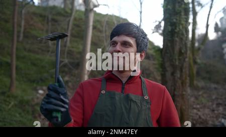Fermier ou jardinier jeune homme en robe rouge avec tablier vert, paire regardant la caméra et souriant, montrant son concept de Hoe , fermier et Jardinage Banque D'Images