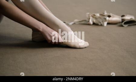 La ballerine est assise sur le sol au studio de danse et met des chaussures pointe avant la leçon de chorégraphie. Danseuse de ballet gracieuse se prépare à la performance ou à l'entraînement. École de danse classique. Pieds rapprochés. Banque D'Images