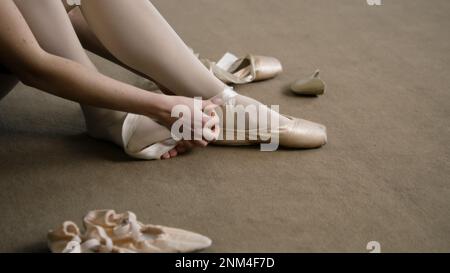 La ballerine est assise sur le sol au studio de danse et met des chaussures pointe avant la leçon de chorégraphie. Danseuse de ballet gracieuse se prépare à la performance ou à l'entraînement. École de danse classique. Pieds rapprochés. Banque D'Images