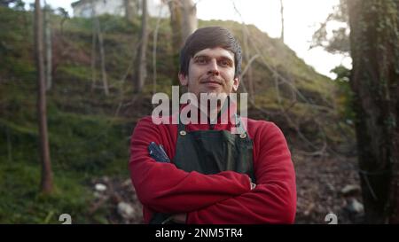 Fermier ou jardinier jeune homme en robe rouge avec tablier vert souriant regardant la caméra et le croisement des bras, Farmer et Gardener concept Banque D'Images