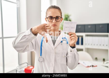 Jeune médecin femme tenant un certificat de covid avec le visage en colère, signe négatif montrant l'aversion avec les pouces vers le bas, concept de rejet Banque D'Images