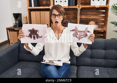 Brunette femme travaillant avec le test de rorschach à la clinique de psychologie peur et choqué avec la surprise et l'expression stupéfaite, la peur et le visage excité. Banque D'Images