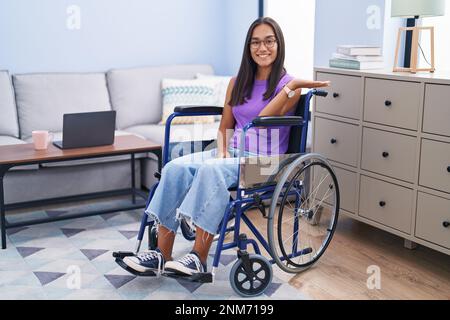 Jeune femme hispanique assise sur un fauteuil roulant à la maison souriante gaie présentant et pointant avec la paume de la main regardant la caméra. Banque D'Images