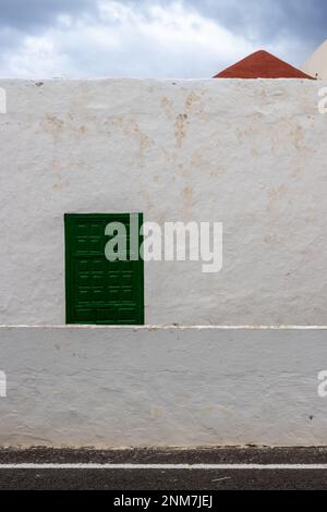 Façade blanche d'une maison à surface rugueuse. Fenêtre avec obturateur fermé vert. Triangle d'un toit rouge vif. Montagne en arrière-plan. Ciel nuageux. A Banque D'Images