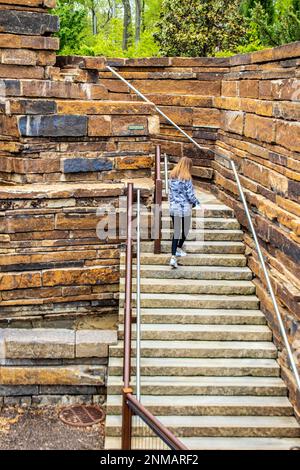 Fille avec de longs cheveux rouges et veste de camouflage monte les marches de roche dans la grande structure de sandstrone avec les arbres et le feuillage au-dessus Banque D'Images