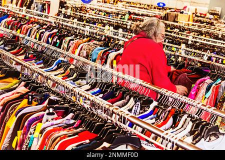 Femme âgée en blouson rouge traversant les porte-vêtements usagés à une rive thrift (bonne volonté) à Ludington, MI, États-Unis. Banque D'Images